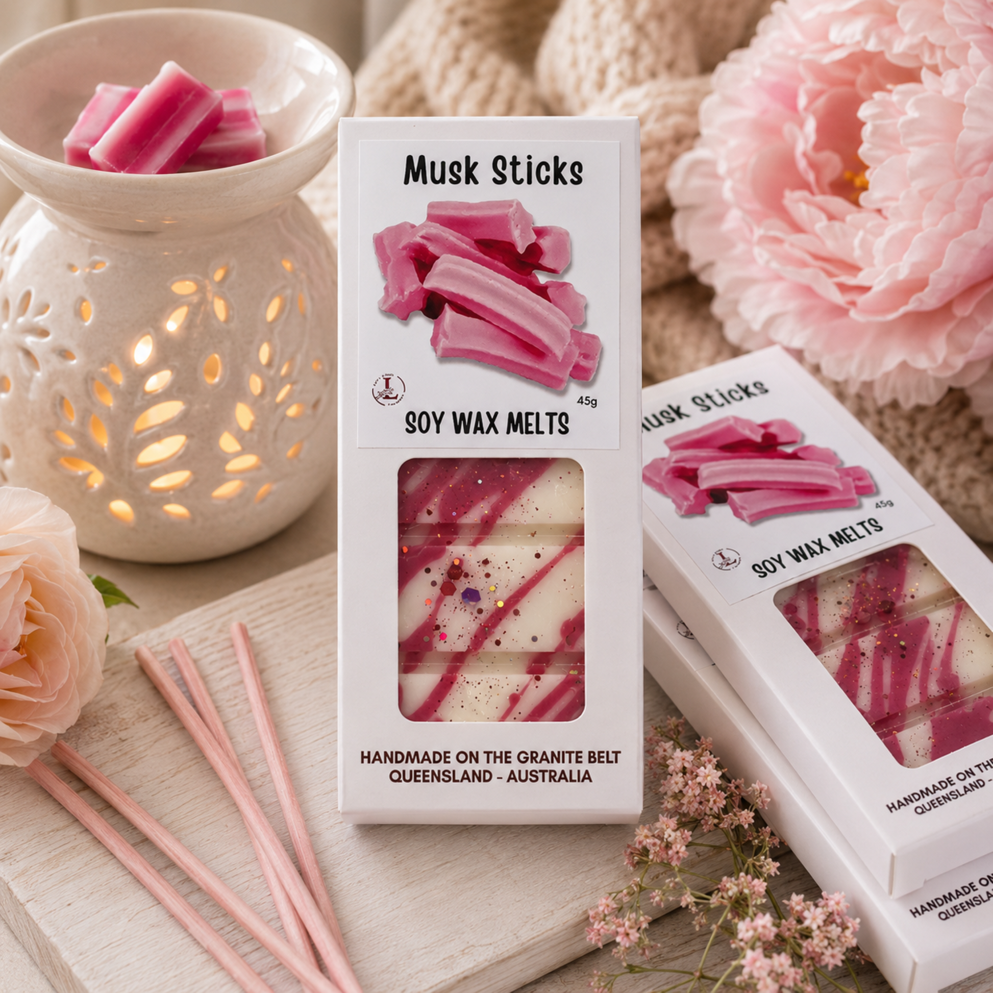 Handmade Musk Sticks premium soy wax melts by Lee's Little Luxuries in a 45g gift box featuring classic pink musk stick candy artwork, displayed beside a glowing ceramic wax warmer, soft blush flowers and pastel décor. Australian-made natural soy wax melts with the nostalgic sweet musk stick fragrance, perfect for creating a warm, cosy and inviting home atmosphere.
