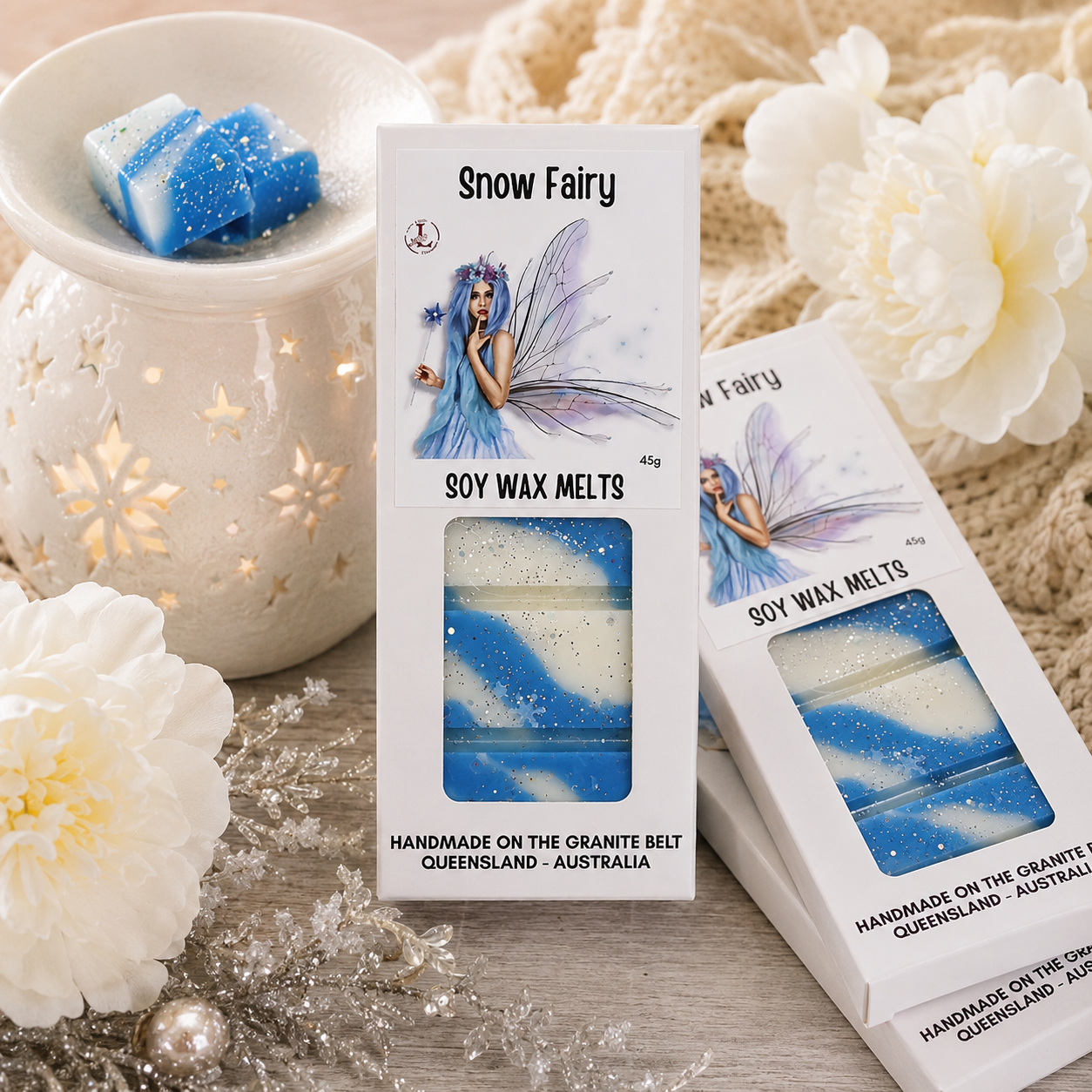 Handmade Snow Fairy soy wax melts by Lee's Little Luxuries in a 45g gift box featuring a whimsical blue fairy design, displayed beside a glowing ceramic wax warmer with blue and white wax melts, soft white flowers and winter-inspired décor. Australian-made premium natural soy wax melts with a sweet, magical fragrance, perfect for creating a cosy and luxurious home atmosphere.