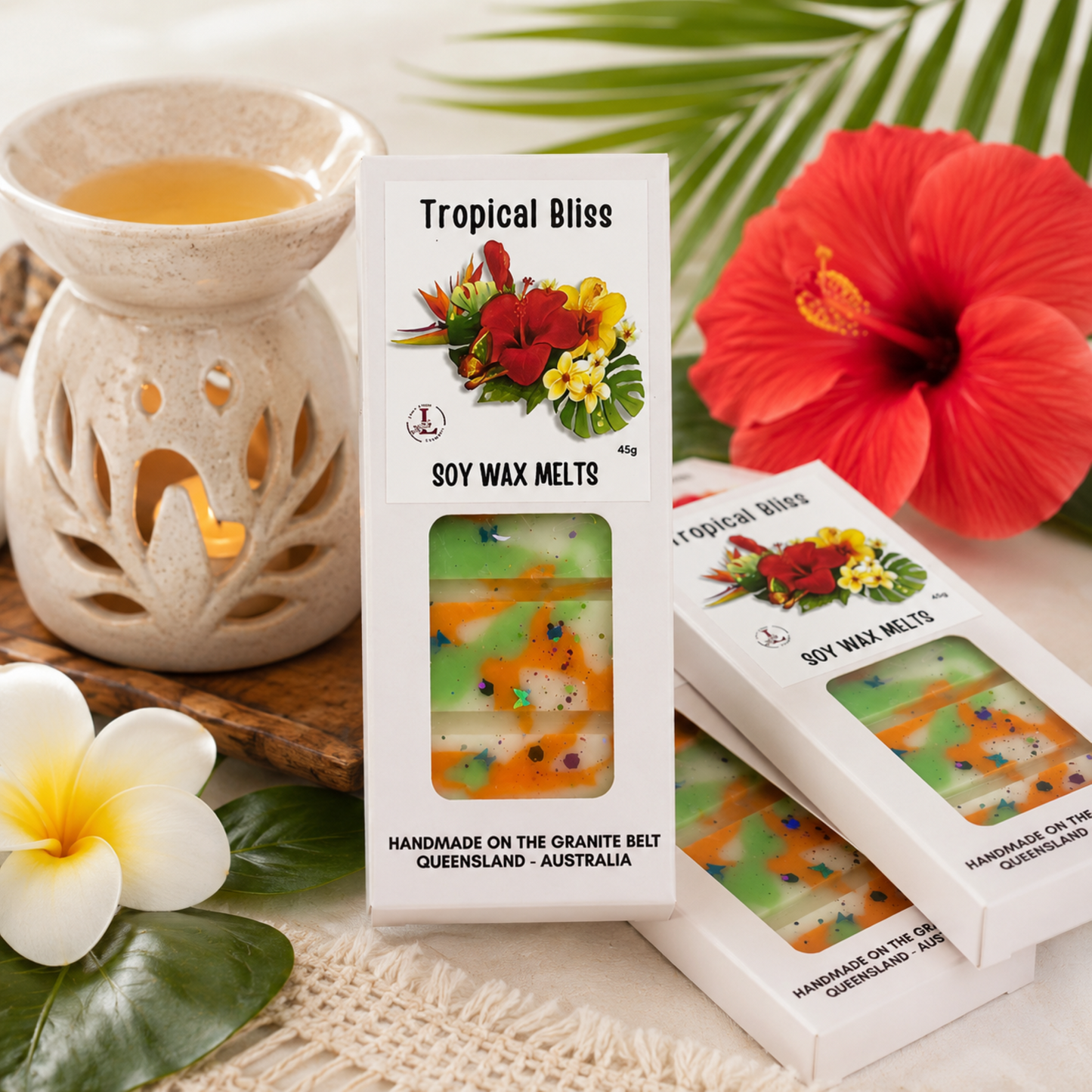 Handmade Tropical Bliss soy wax melts by Lee's Little Luxuries in a 45g gift box with tropical hibiscus flower artwork, displayed beside a ceramic wax warmer, tropical flowers and palm leaves. Australian-made natural soy wax melts with a fresh tropical fragrance, ideal for home fragrance and gifts.